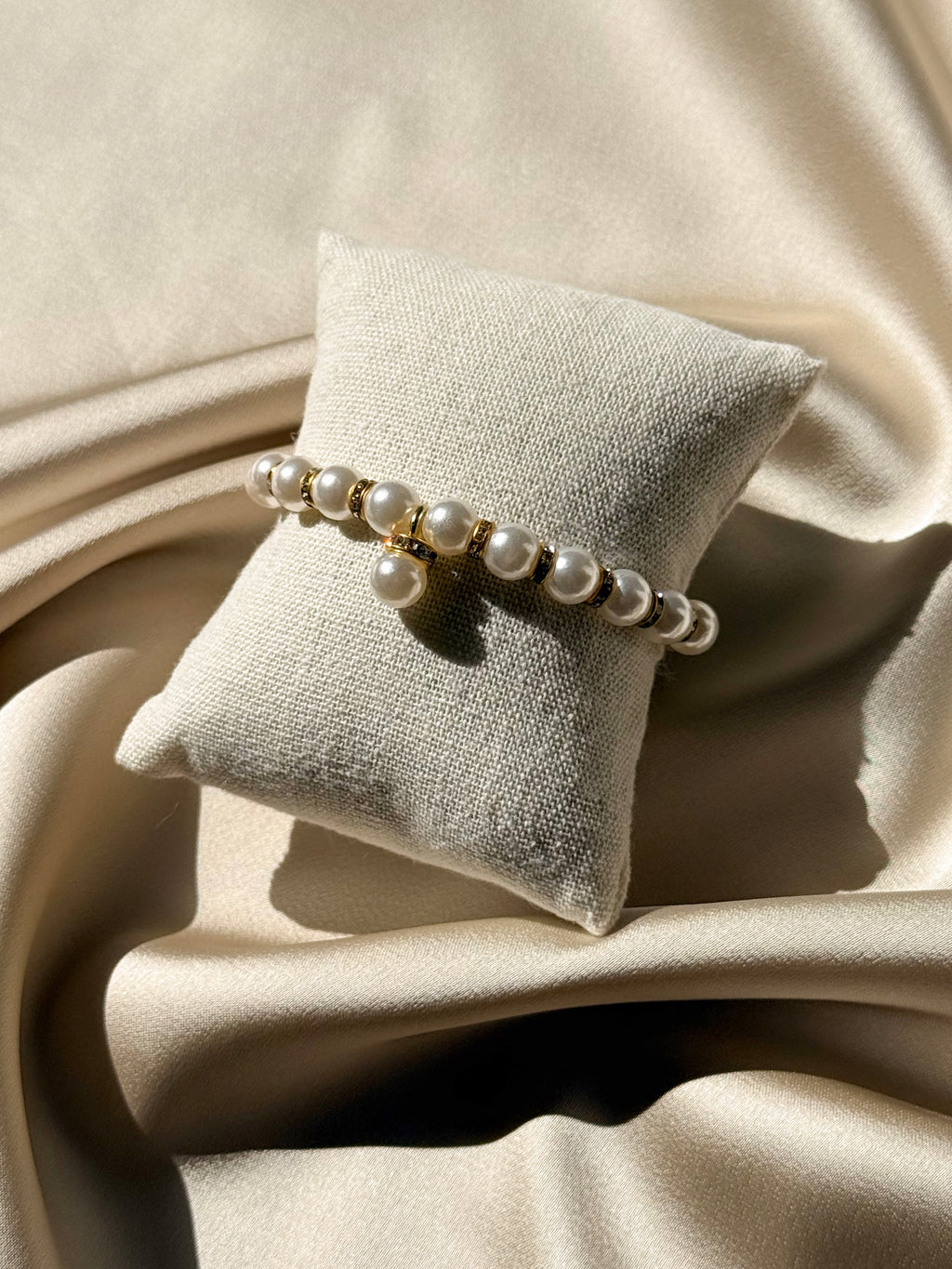 A gold-toned beaded bracelet with white faux pearls draped over a small beige fabric jewelry pillow, set against a backdrop of draped satin fabric.