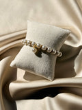 A gold-toned beaded bracelet with white faux pearls draped over a small beige fabric jewelry pillow, set against a backdrop of draped satin fabric.