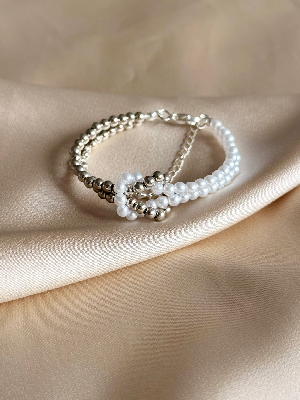 A double-strand beaded bracelet featuring white faux pearls and polished silver-tone beads, displayed on draped beige satin fabric.