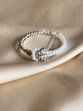 A double-strand beaded bracelet featuring white faux pearls and polished silver-tone beads, displayed on draped beige satin fabric.