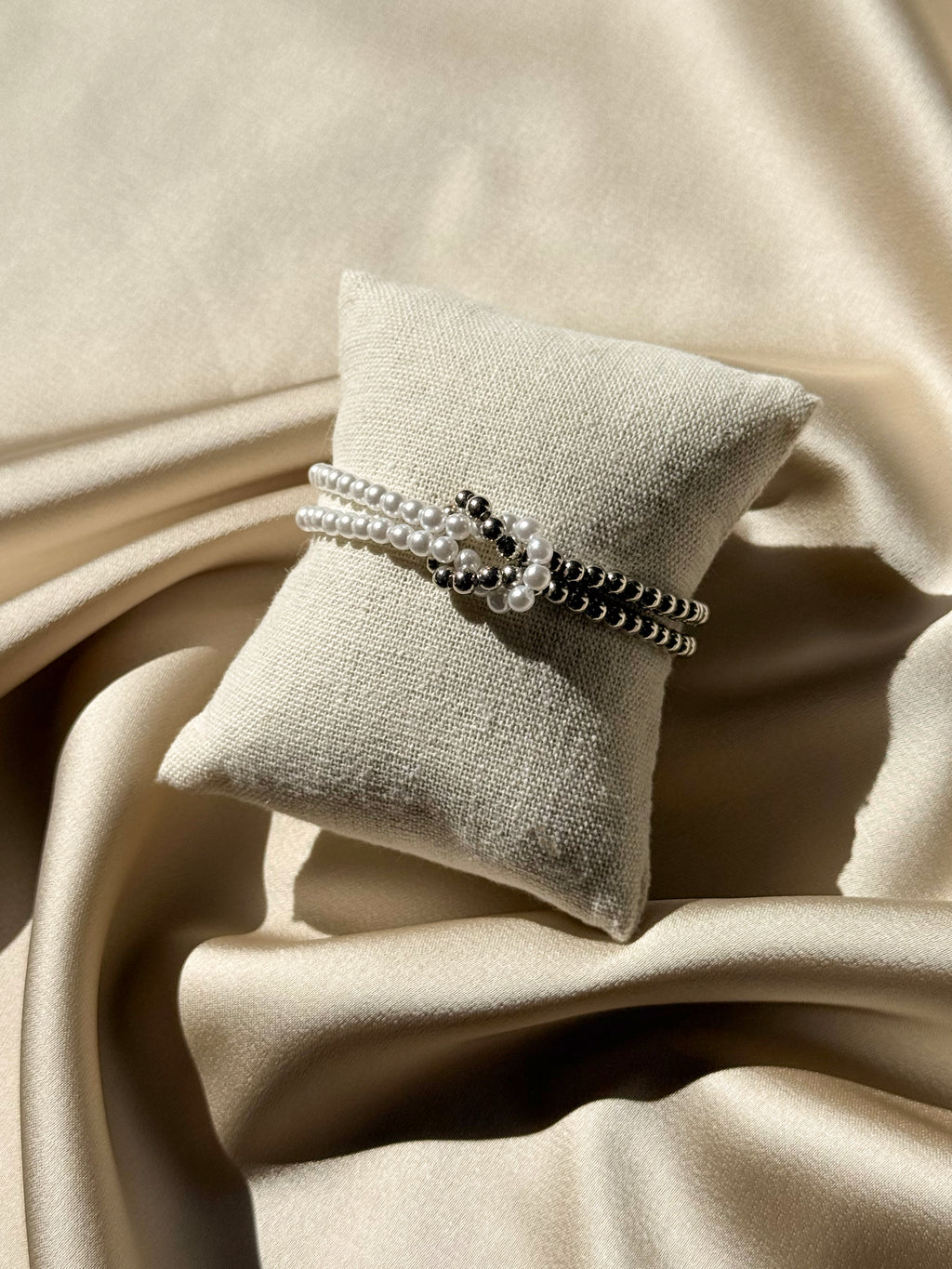 A beaded wrap bracelet with white pearl-like strands and black seed beads, displayed on a small beige fabric pillow over draped champagne satin fabric.