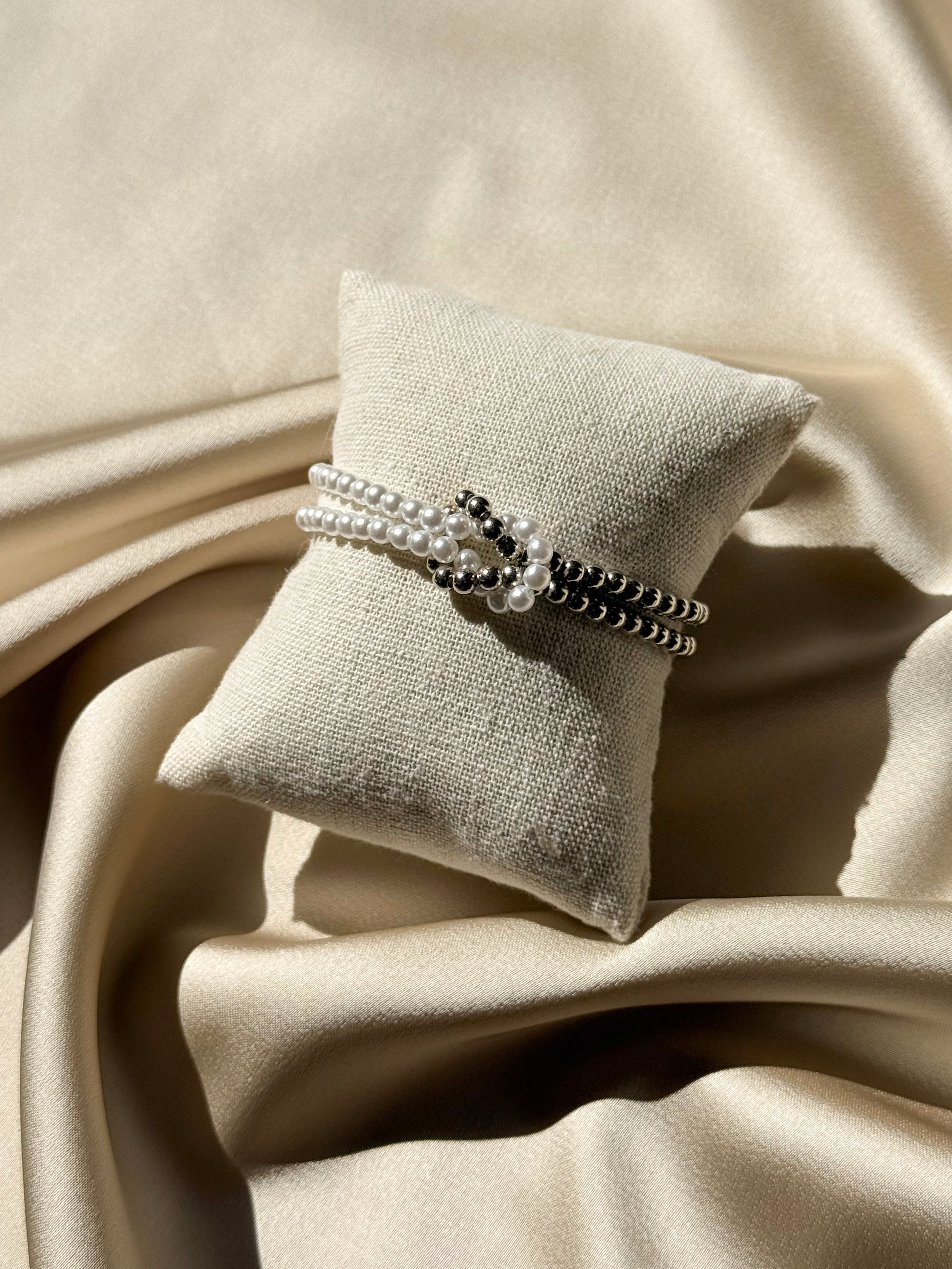 A beaded wrap bracelet with white pearl-like strands and black seed beads, displayed on a small beige fabric pillow over draped champagne satin fabric.