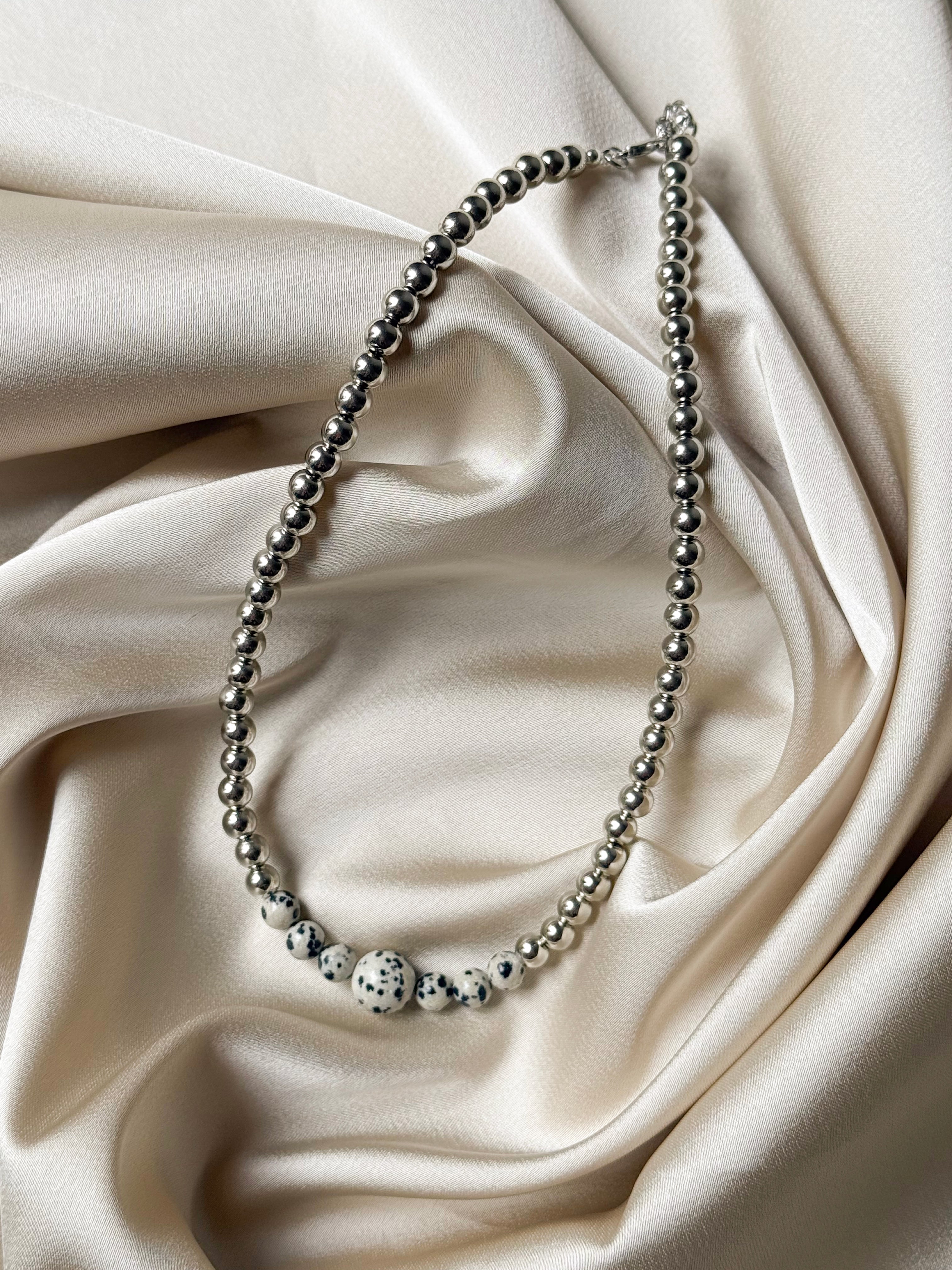 A silver beaded necklace with a central white and black spotted bead, displayed on draped cream satin fabric.