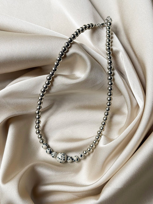 A silver beaded necklace with a central white and black spotted bead, displayed on draped cream satin fabric.