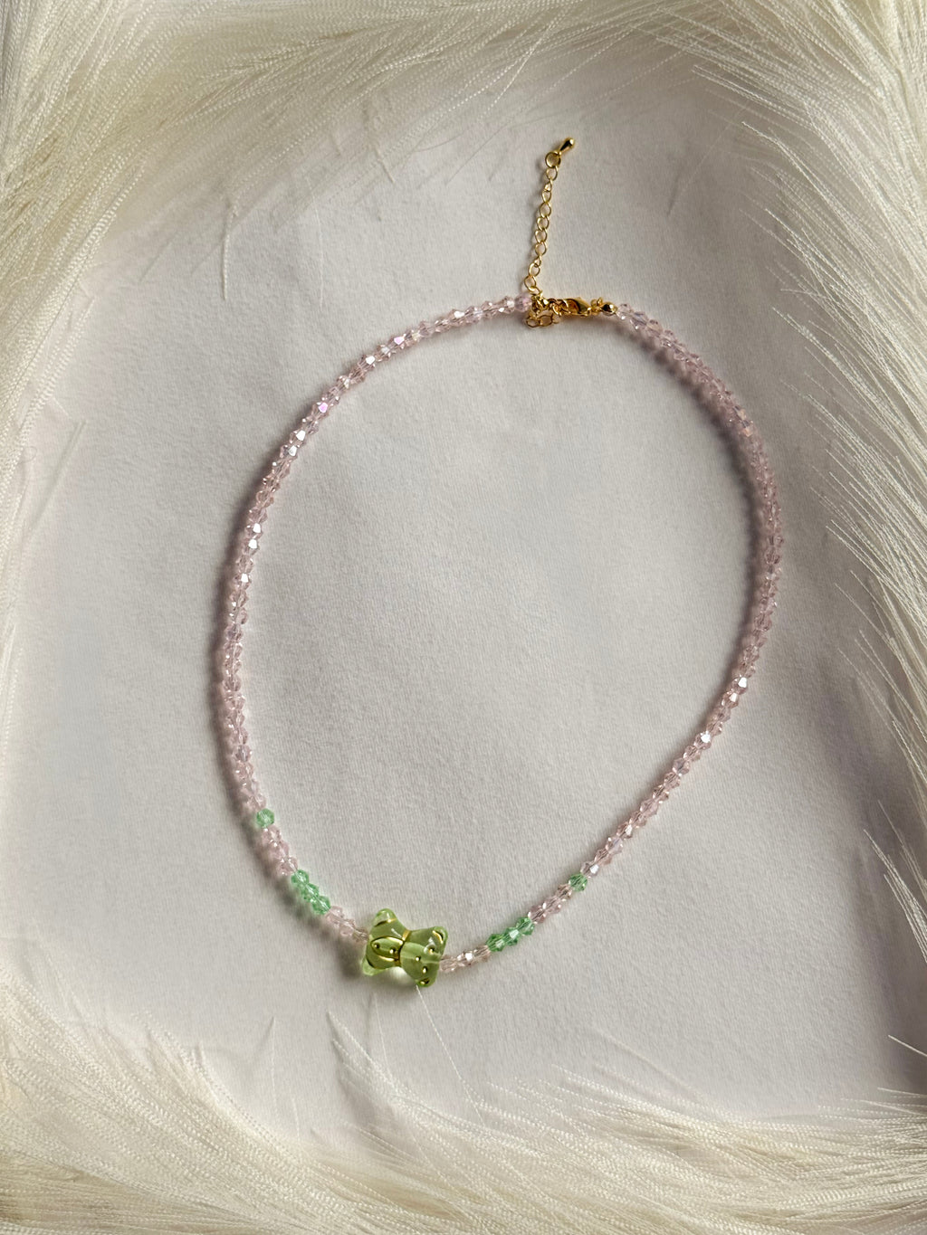 A delicate beaded necklace with light pink beads, small green accents, and a central green bow charm, displayed on a soft white faux fur background.