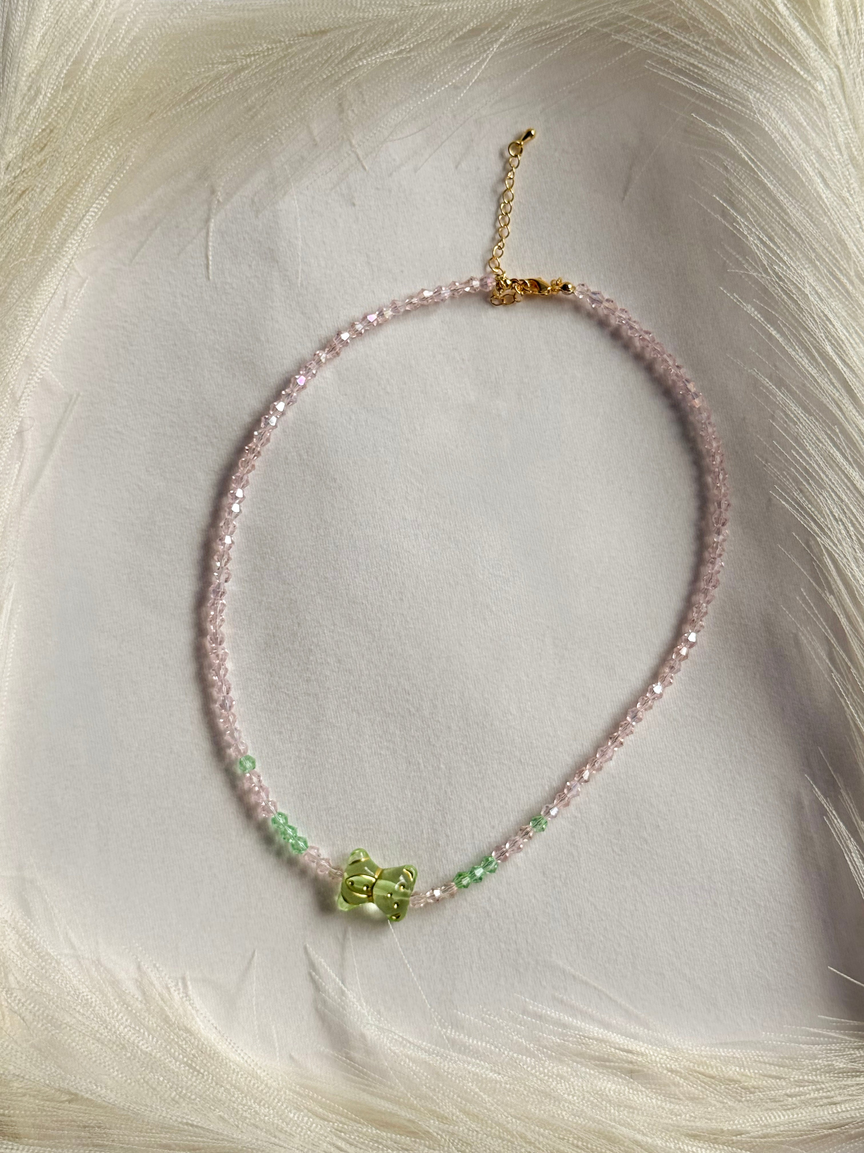 A delicate beaded necklace with light pink beads, small green accents, and a central green bow charm, displayed on a soft white faux fur background.
