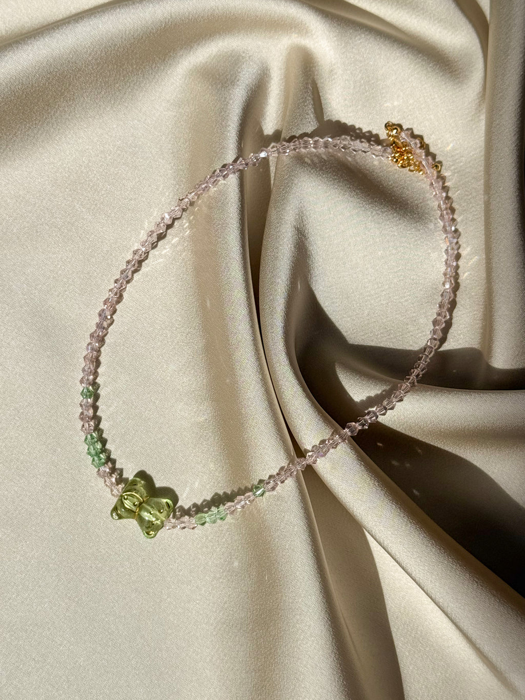 A delicate beaded necklace with light pink and green beads, featuring a small green butterfly charm and a gold-tone lobster clasp, displayed on draped cream satin fabric.