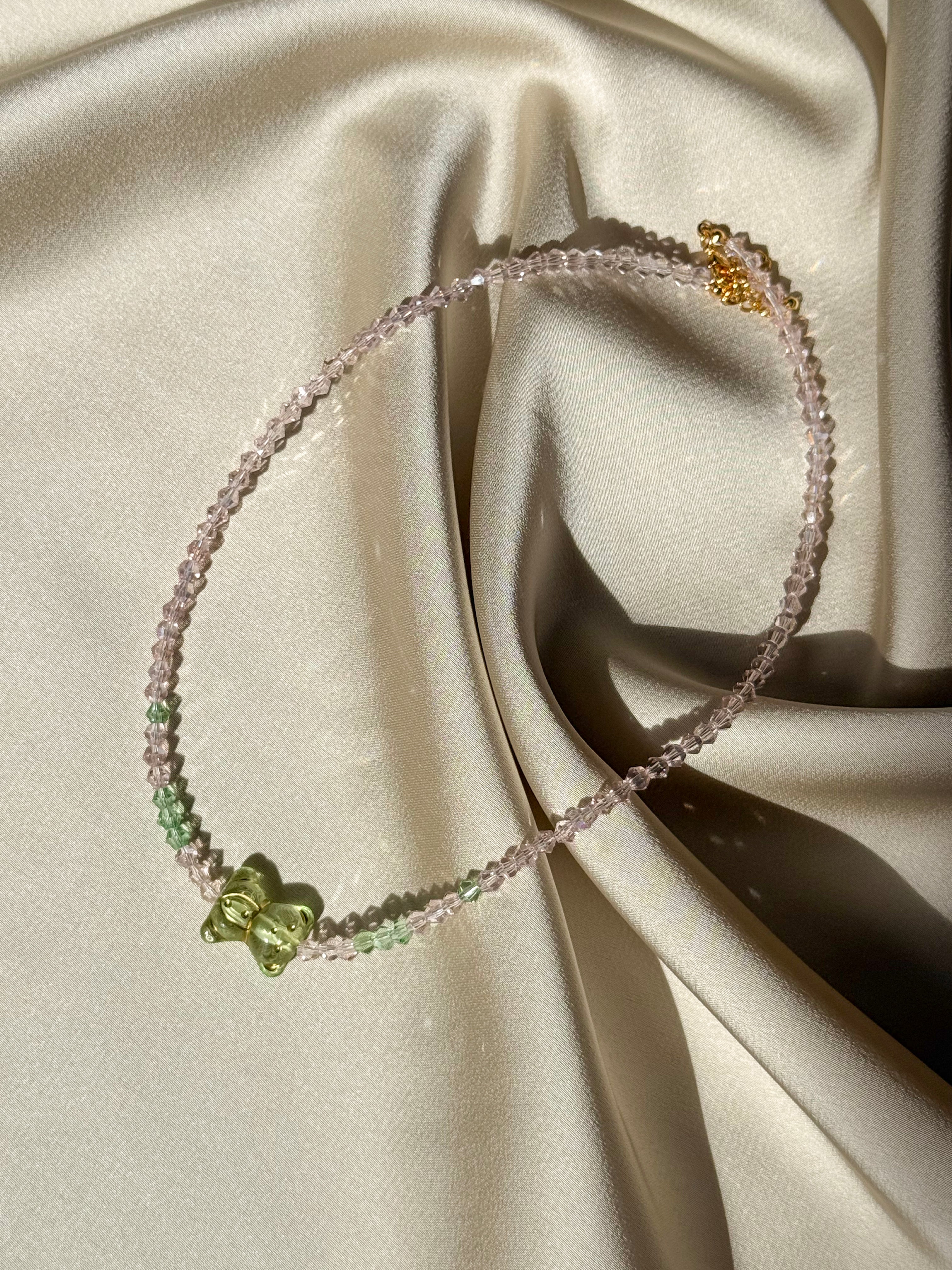 A delicate beaded necklace with light pink and green beads, featuring a small green butterfly charm and a gold-tone lobster clasp, displayed on draped cream satin fabric.